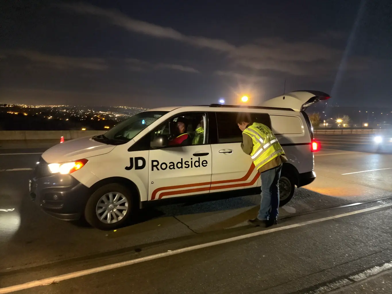 An image showing a RoadAid service vehicle jump-starting a car in a parking lot. The scene is set at night, with the car's headlights illuminating the area, emphasizing RoadAid's 24/7 availability.